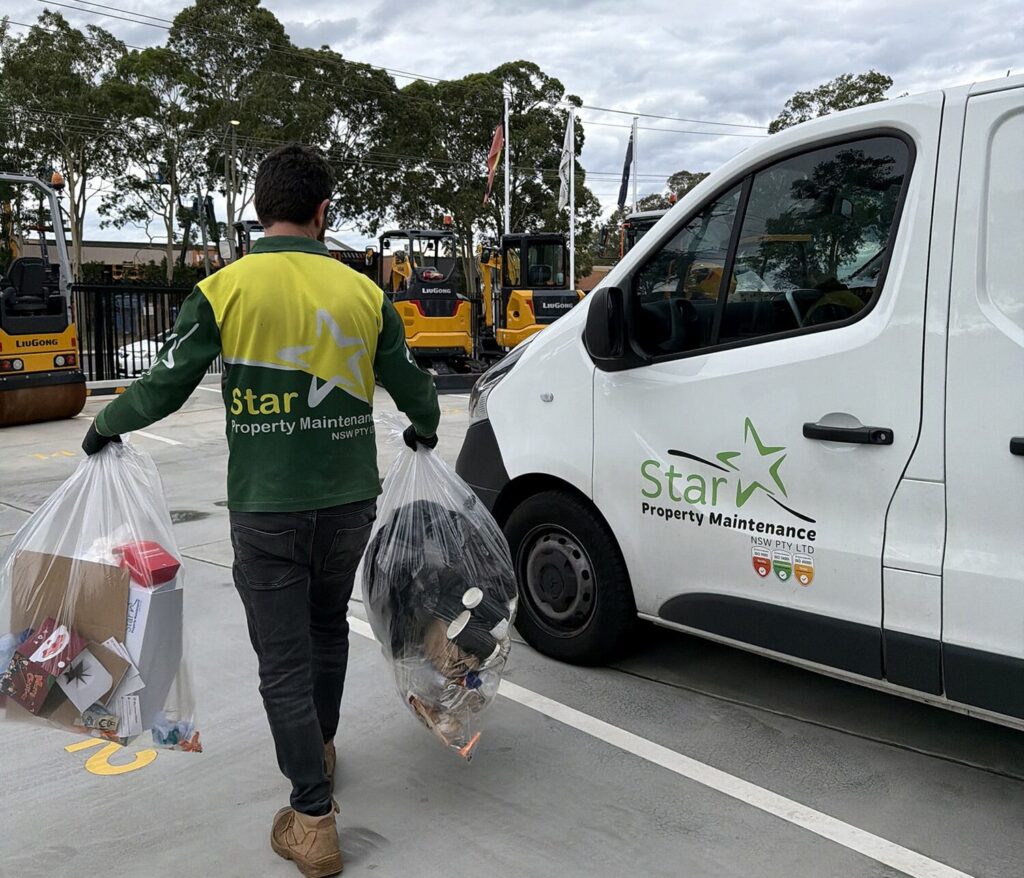 Worker in green and yellow hi-vis shirt carries two clear rubbish bags past a white work van in a car park.